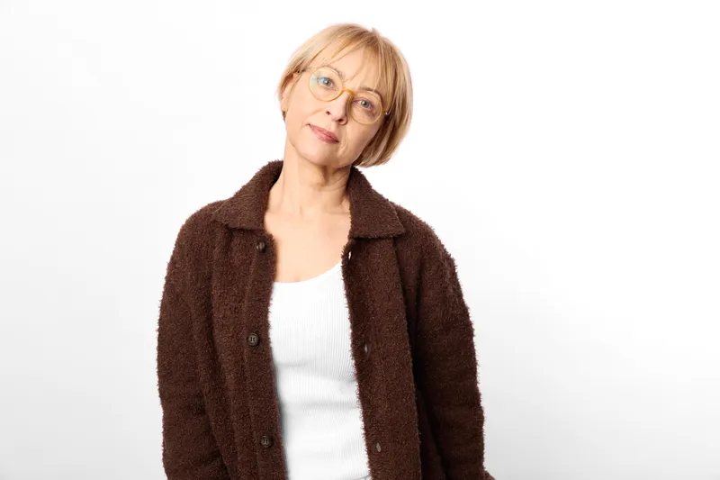 Elderly woman with glasses, brown jacket, surprised expression on a clean white background, close studio portrait showing aging and concern, soft lighting and neutral mood.