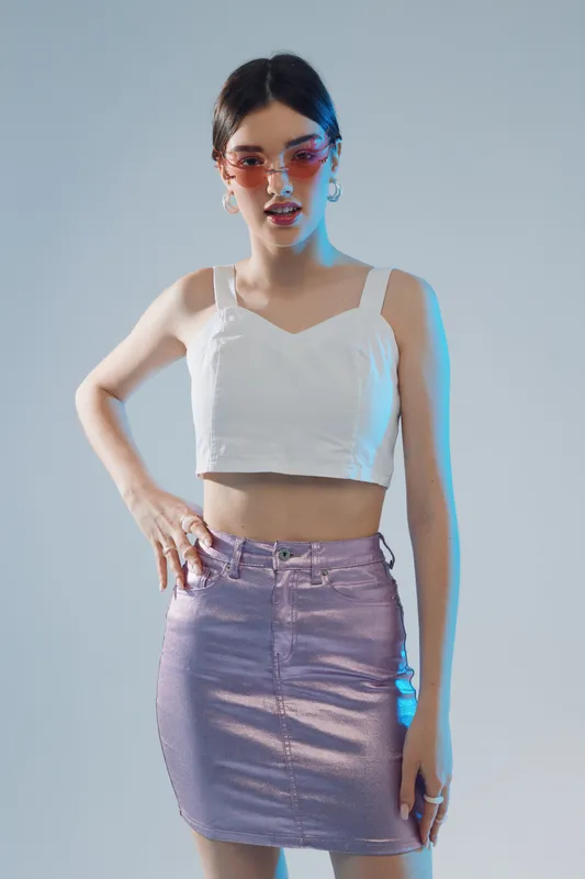 Young woman in stylish sunglasses wearing a white crop top and purple skirt poses confidently against a neutral background. Concept of woman beauty fashion.