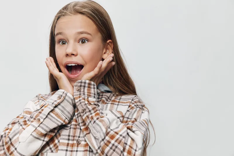 Girl with long hair expressing surprise and excitement with open mouth and hands on cheeks wearing plaid shirt on light background.