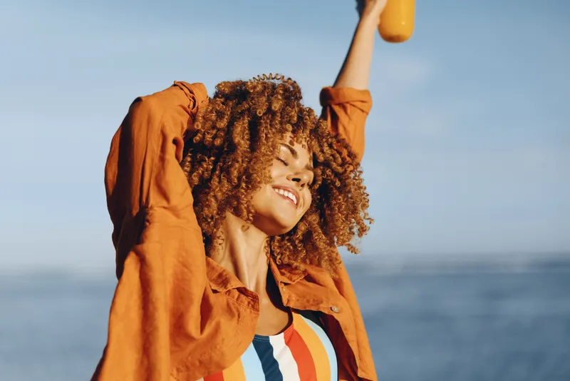 Child with curly hair wearing an orange jacket smiles by the ocean arms raised holding a yellow balloon carefree summer outdoors freedom and happiness on sunlit beach with blue sky horizon