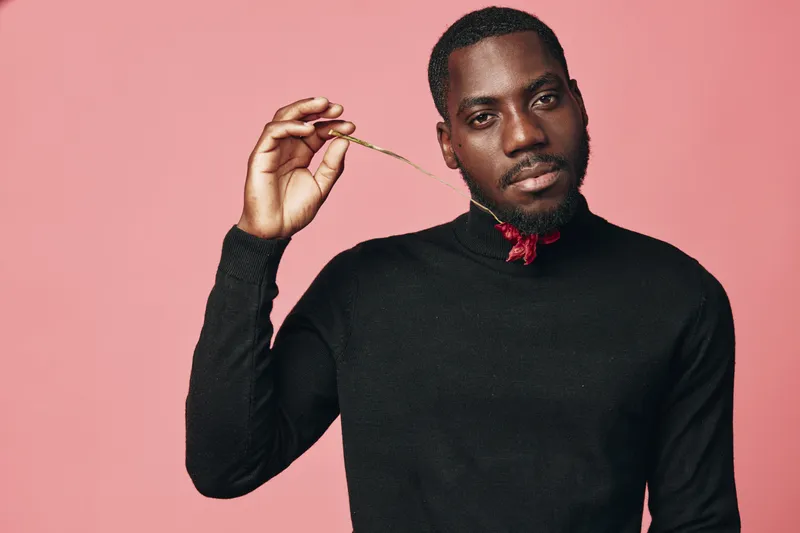African man with dark skin wearing black sweater and red bow tie holding gold chain against pink background in creative conceptual visual style portrait for fashion and elegance.