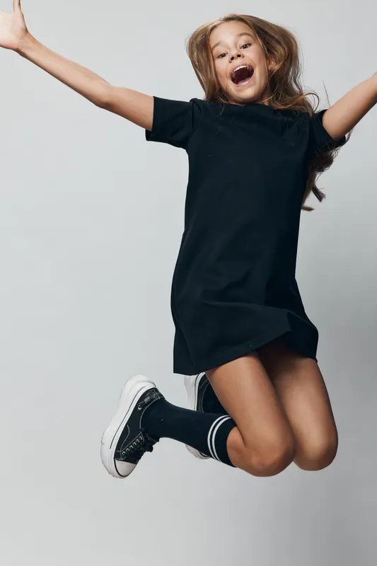Girl jumping with joy in black dress and sneakers, smiling with open arms against gray background in studio for happiness and energy concept.