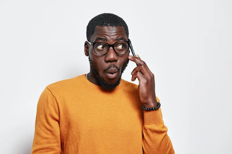 Black man with dark skin wearing glasses and orange sweater talking on smartphone with surprised expression on white background. African ethnicity, communication and technology concept.