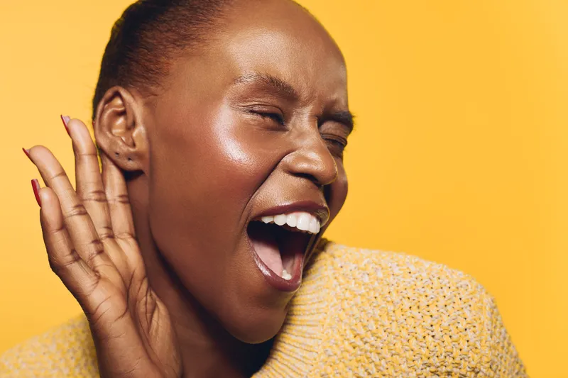 Excited dark skinned African woman in yellow sweater laughs with eyes closed, hand near ear. Studio portrait on yellow background. Concept for joy, happiness, and positive emotions.