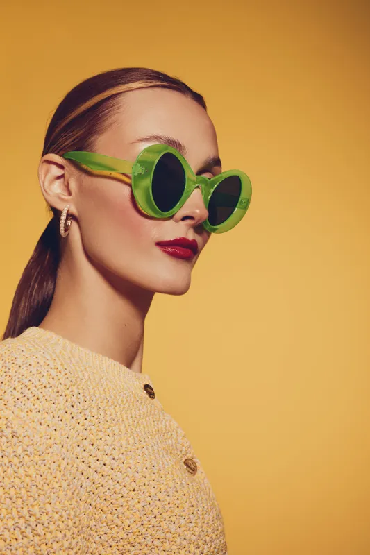 Stylish woman wearing green round sunglasses and yellow knitted sweater poses on yellow background. Fashion portrait with bright colors and modern accessories for trendy look.