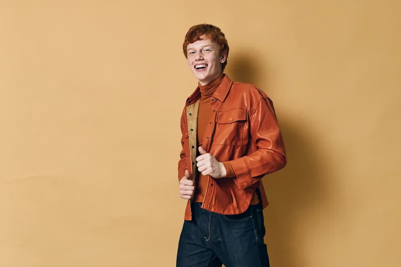 Young man with red hair wearing orange jacket and brown turtleneck smiling and posing against beige background in studio portrait with casual fashion style.