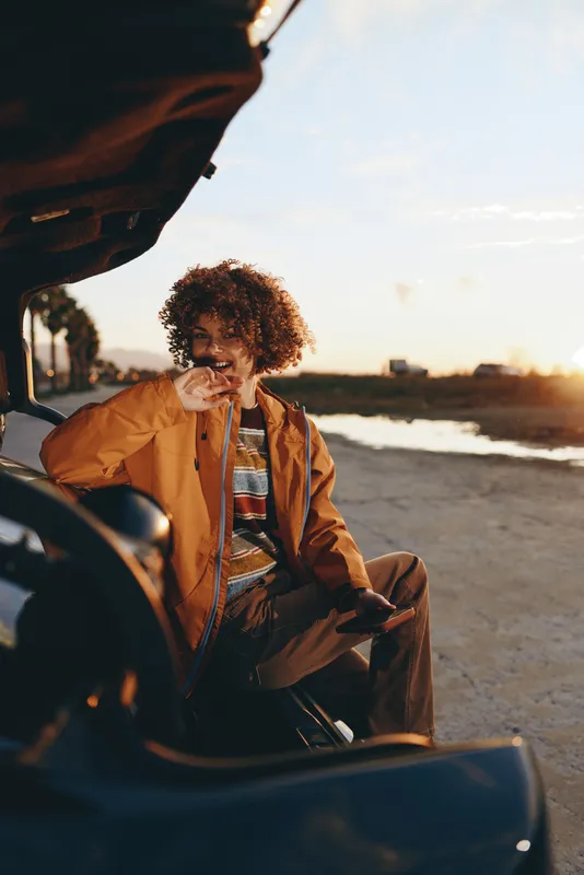Woman lifestyle at the beach during sunset, curly hair visible as she sits beside a car, wearing a warm jacket, holding a small dog, coastal golden light, travel mood
