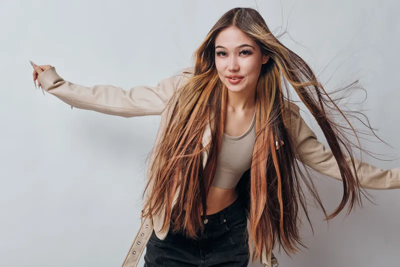 Young woman with long straight hair wearing beige top and cardigan moving with raised arms against white background with confident and cheerful expression smiling.