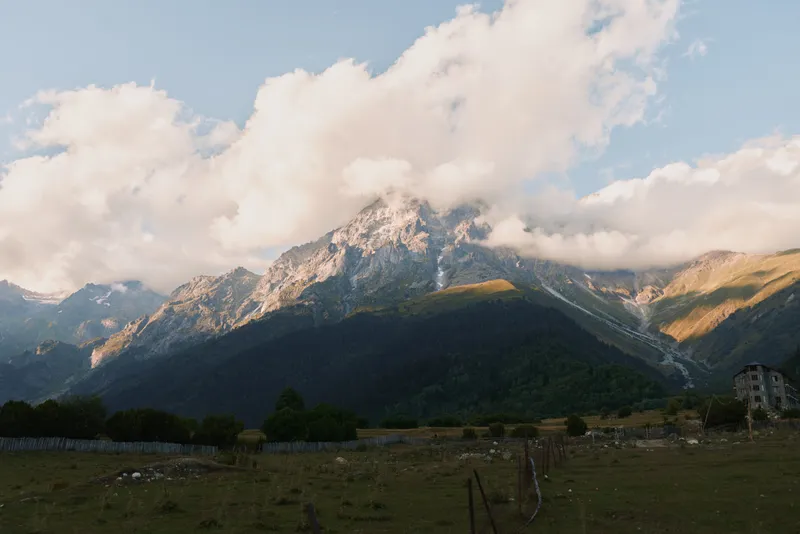 Sunlit alpine landscape with towering mountains, a wooden fence in the foreground and distant houses creating a peaceful rural scene.