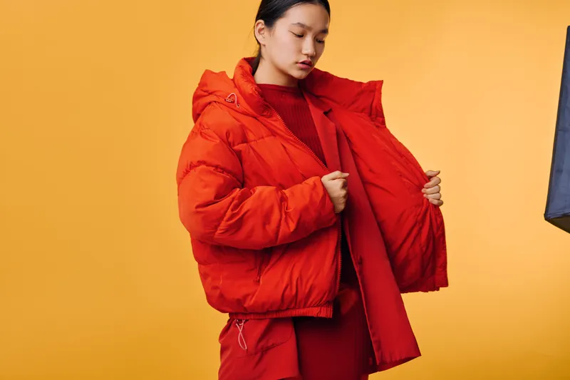 Woman wearing bright orange puffer jacket and matching outfit standing against yellow background with closed eyes and holding jacket open for display with thoughtful expression.