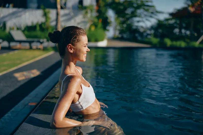 A woman relaxes at the pool edge, shoulders above water, enjoying warm sunlight. A lifestyle moment that conveys leisure, calm, and wellness in a bright outdoor setting.