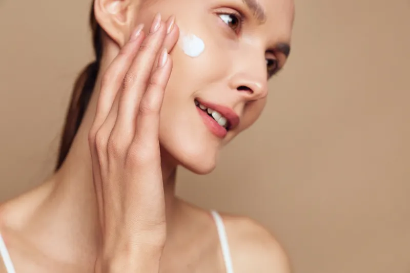 Young woman with long brunette hair applies cream on her cheek while smiling gently against a neutral background in a close-up skincare routine.
