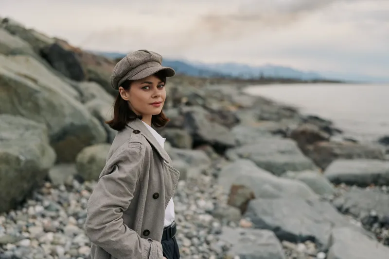 Young woman in vintage coat and cap stands on rocky beach looking at camera. Overcast sky and sea in background. Concept for fashion, autumn, and outdoor portrait.