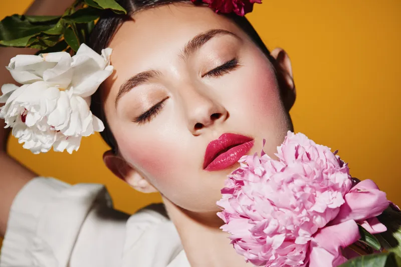 Close-up portrait of a woman with creative fashion makeup wearing floral headpiece holding pink peony. Concept for beauty, creativity, summer look, and feminine styling with yellow background.