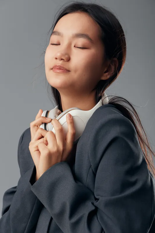 Young woman with closed eyes holding headphones near her chest in gray jacket, enjoying music and peaceful moment with calm expression on neutral background for relaxation and mindfulness.