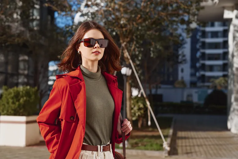 A stylish woman in a red coat and green turtleneck walks outdoors in an urban plaza, exuding confidence as she glances to the side under bright daylight.