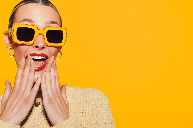 Surprised young woman with yellow sunglasses and headband covers mouth with hands on bright yellow background. Concept of excitement, fashion, and vibrant summer style.