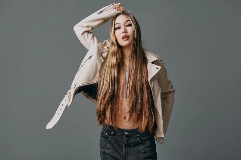 Young woman with long hair wearing open leather jacket and jeans posing confidently against grey background in fashion portrait studio shot.