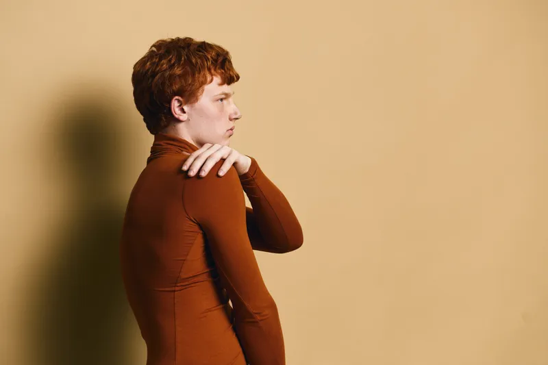 Young man with red hair and freckles wearing brown fitted long sleeve shirt standing sideways against beige background with thoughtful expression and hand on shoulder.