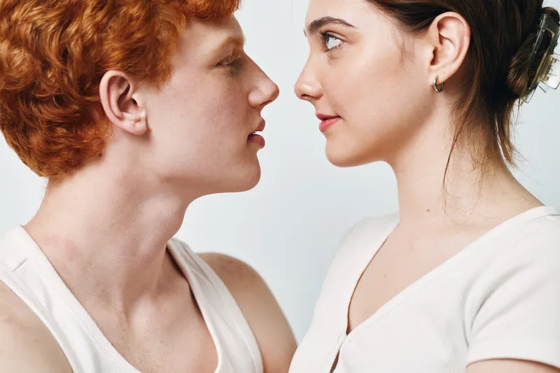 Young woman and man face to face in closeup, looking at each other with neutral expression. Intimate moment with minimalistic white background and natural lighting.