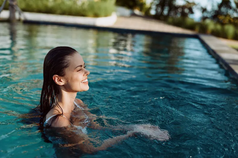 A smiling woman enjoys swimming in an outdoor pool on a sunny day surrounded by greenery. The lifestyle image shows relaxation and happiness in a natural setting.