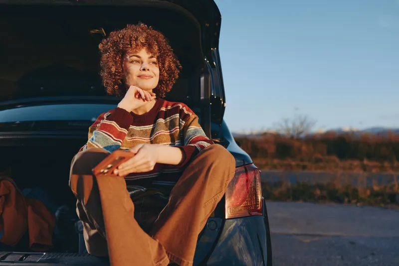 Woman lifestyle, sitting in open car at golden sunset, curly hair softly blown by breeze, warm smile, relaxed posture, cozy sweater, evening road background, natural colors, calm mood