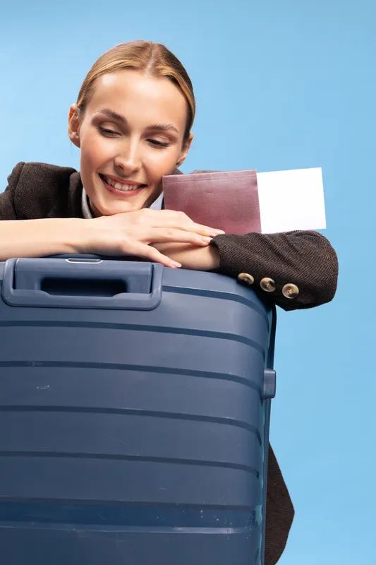 Smiling woman leans on large blue suitcase holding passport and ticket, ready for travel. Studio portrait on blue background. Concept of vacation, tourism, and trip preparation.