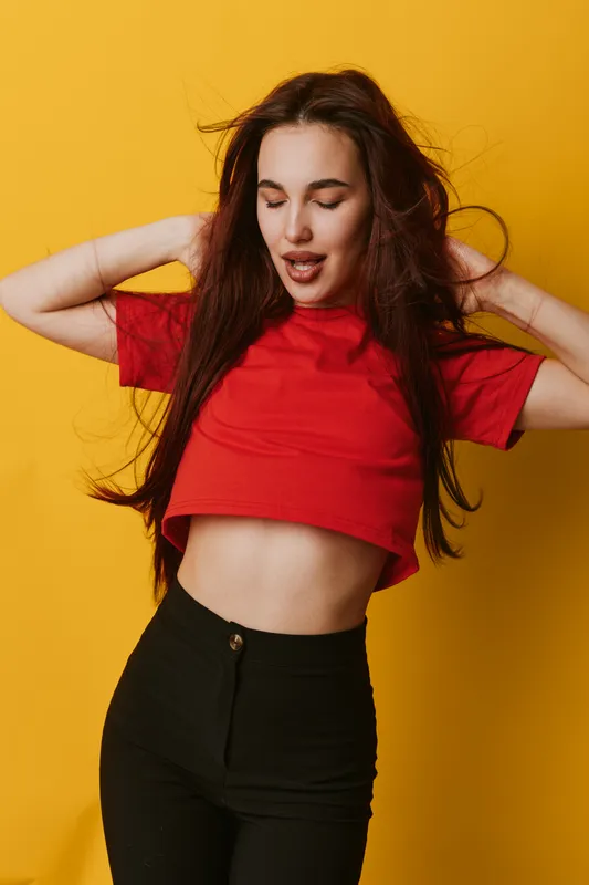 Young woman posing with eyes closed and hands behind head wearing red crop top and black pants against yellow background. Casual style and relaxed mood with long hair flowing.