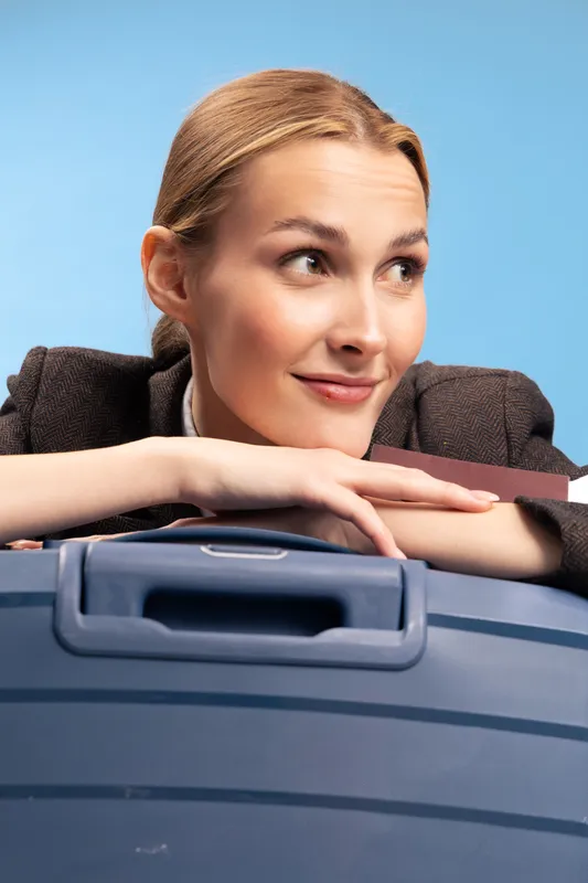 Young businesswoman with light hair rests on a blue suitcase, looking to the side with a slight smile. Concept of travel, business trip, and positive anticipation.