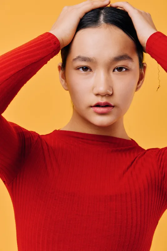 Young woman in red sweater posing with hands on head against orange background. Closeup portrait of female with natural look, dark hair, and focused facial expression in studio lighting.