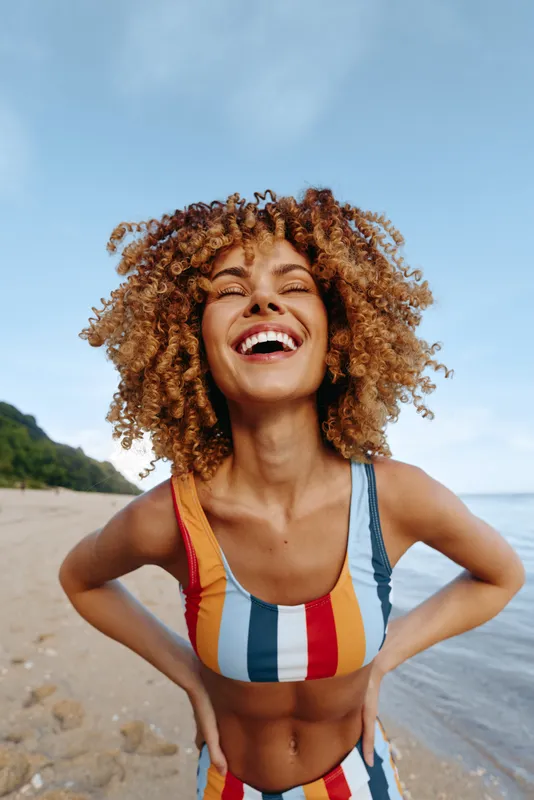 Joyful woman with curly hair wearing a striped swimsuit on a sunny beach, posing with hands on hips, bright smile, carefree summer mood, seaside scenery.