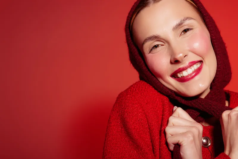 Smiling woman in red coat and scarf poses against red background. Close-up portrait with bright lipstick and natural skin glow. Concept of fashion, winter style, and happiness.