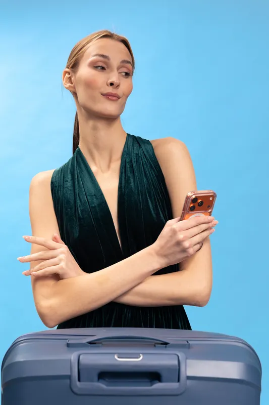Confident woman in elegant dress holding smartphone stands behind suitcase on blue background. Concept of travel, communication, and stylish female traveler waiting for trip or journey.