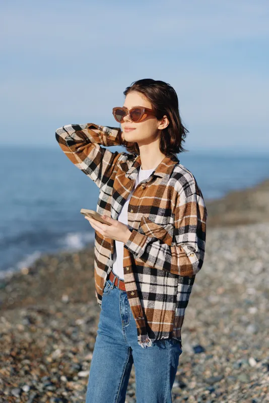 Woman on a sunny beach wearing a plaid shirt and sunglasses, holding a smartphone, gazing toward the ocean in a casual coastal portrait.