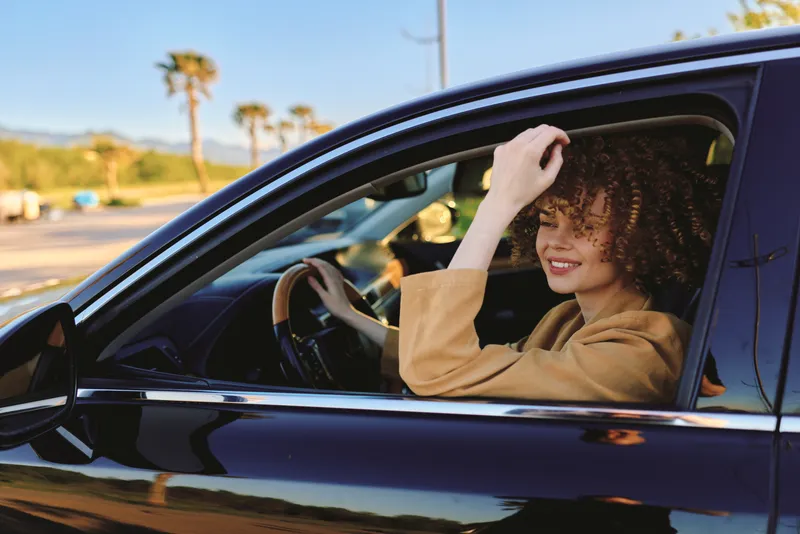 Young woman smiles while driving a black car in a sunlit street, captured with warm retro film color and a carefree vibe.
