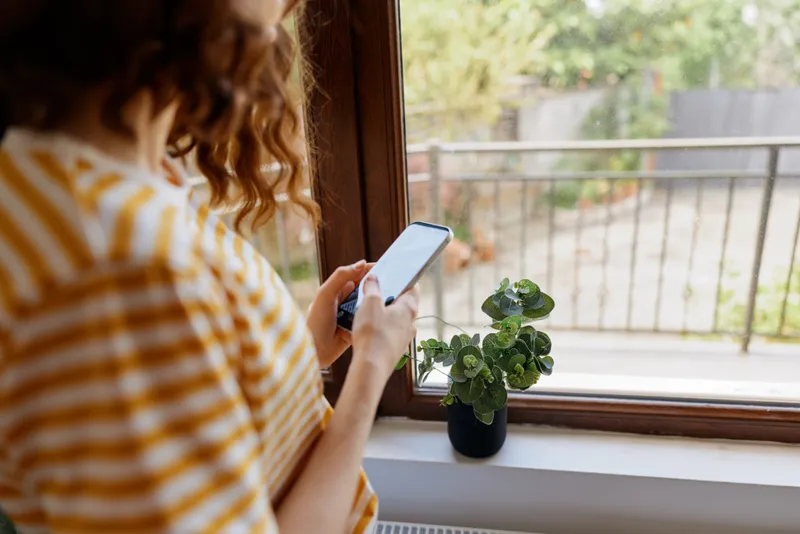 Young woman with a striped yellow shirt sits by a sunlit window and checks her smartphone, a small potted plant on the sill, creating a calm home lifestyle moment. Soft morning light adds warmth, a