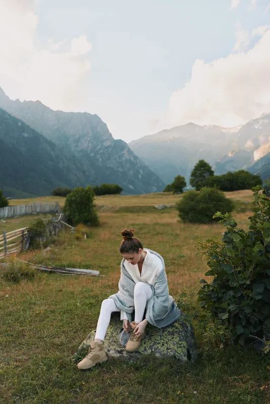 Woman sitting in a meadow near mountains, wearing a cozy white outfit, enjoying a calm outdoor moment in a natural rural setting, highlighting lifestyle and serenity
