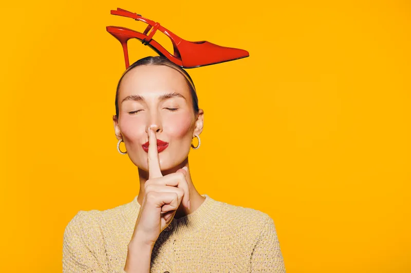 Young woman with closed eyes holds finger to lips in silence gesture. Bright orange background and red high heel shoe on head create playful and stylish concept of quiet and fashion.