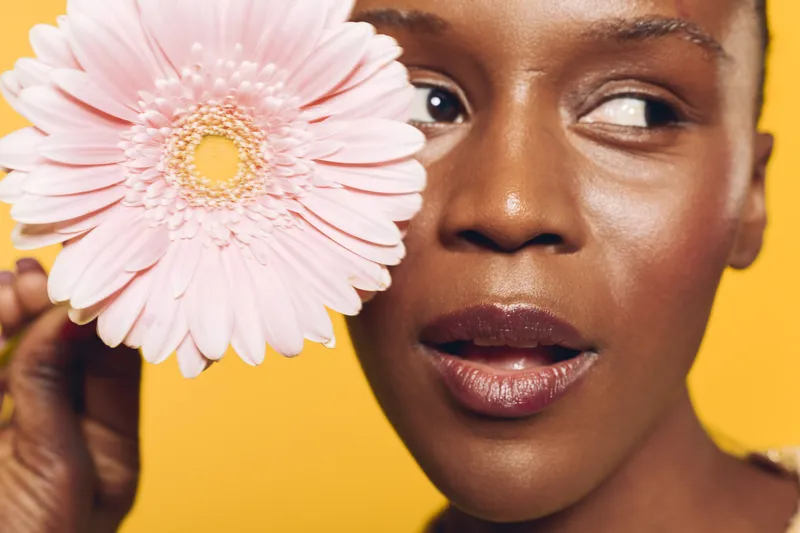 Close-up of smiling dark skinned African woman holding pink flower near face on yellow background. Concept of natural beauty, skincare, wellness, and vibrant healthy lifestyle with floral element.