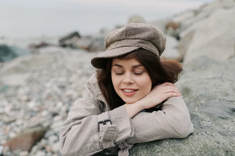 Young woman in beige coat and tweed cap smiles gently with closed eyes resting on stone by rocky beach. Concept of relaxation, nature, autumn, and peaceful moment outdoors.