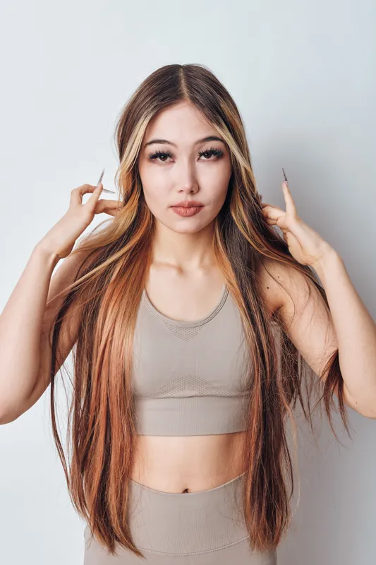 Young woman with long straight hair in beige top posing against white background, looking at camera with calm expression touching hair with fingers indoors.