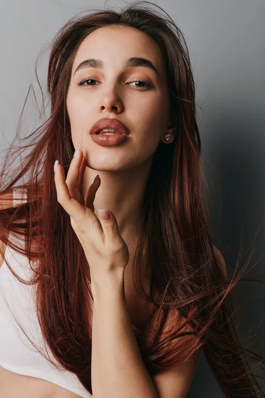 Woman with long hair and natural makeup posing with hand near face against gray background. Beauty portrait with confident expression and casual white top in studio lighting.