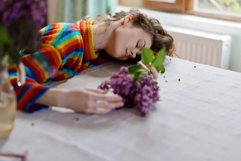 A young woman in a colorful striped sweater leans on a table, resting her head beside a purple bouquet, conveying quiet contemplation and a calm home mood.