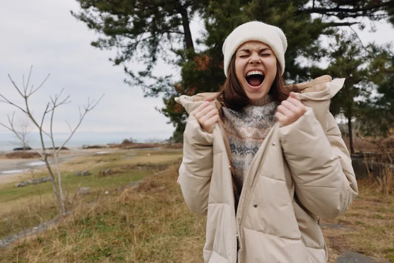 Excited woman in a winter coat and scarf enjoys an open air moment in a natural landscape, wind in the trees and distant water, expressing joy and freedom outdoors