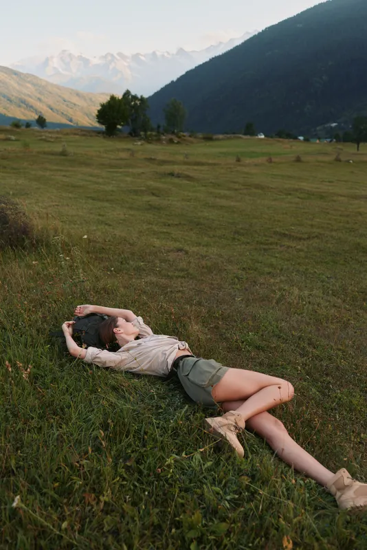 A young woman with brunette hair lies relaxed on green grass in a wide open meadow surrounded by mountains during a calm late afternoon.