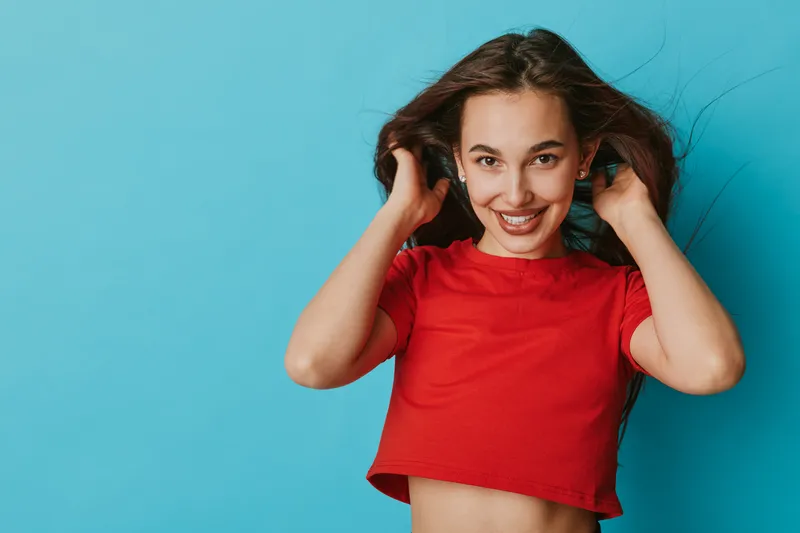 Young woman smiling and playing with hair wearing red crop top against blue background. Portrait of happy girl with long hair and casual style in studio with bright lighting.