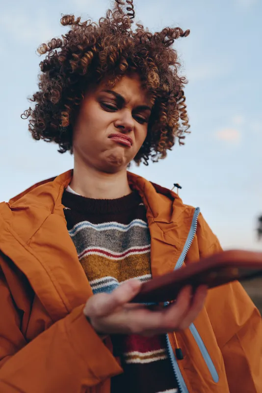 Woman lifestyle portrait, curly hair and orange jacket outdoors, she checks smartphone on phone screen with relaxed calm expression, casual city daylight background, closeup young female reading