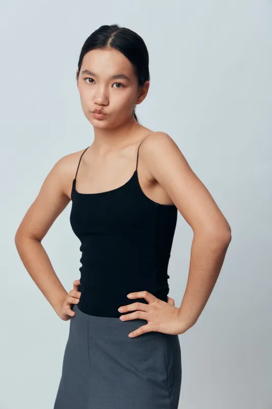 Young woman standing with hands on hips wearing black tank top and gray skirt against plain background confident and serious expression with natural makeup and sleek hairstyle.