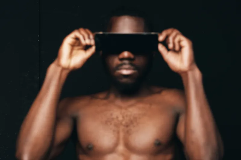 Black man with dark skin holding virtual reality headset on face in creative conceptual visual style with bare chest and blurred background for technology and innovation.