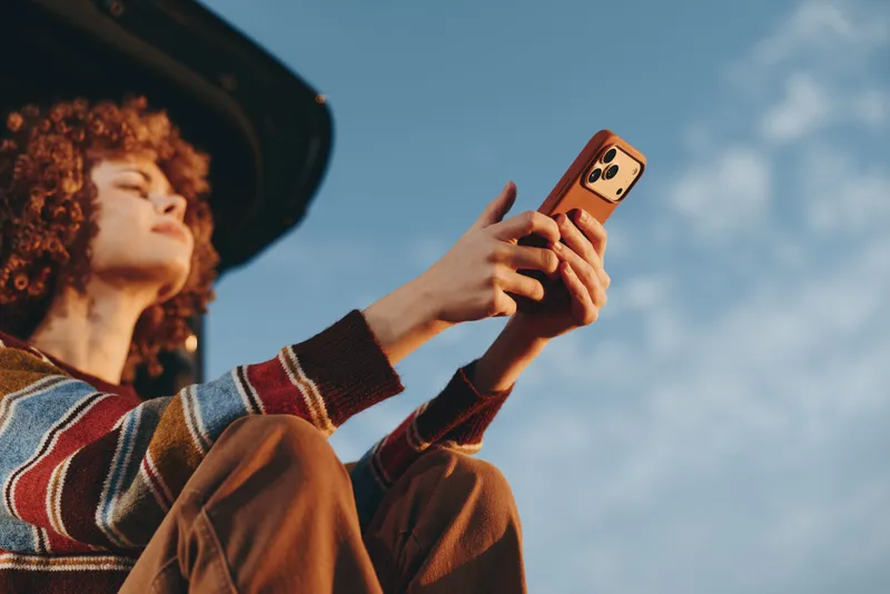 Woman lifestyle outdoors, smiling holding smartphone for selfie, wearing hat and striped sweater, technology moment, summer sky background, relaxed casual mood, soft natural light